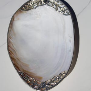 May include: Oval-shaped decorative dish with a mother-of-pearl interior and a silver-colored metal rim. The rim features floral and leaf designs at the top and bottom. The dish has a pearlescent white and brown color scheme.