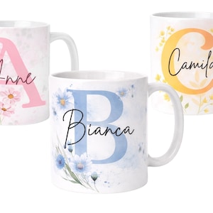 May include: Three white ceramic mugs with floral and initial designs. One mug features a pink "A" with the name "Anne" and pink flowers. Another has a blue "B" with "Bianca" and blue flowers. The third mug displays a yellow "C" with "Camila".