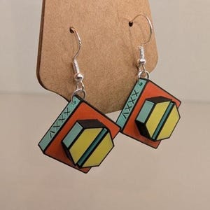May include: A pair of geometric earrings with silver hooks. Each earring features a red square with a light blue border and a yellow and black hexagon in the center. The earrings are displayed on a brown card.