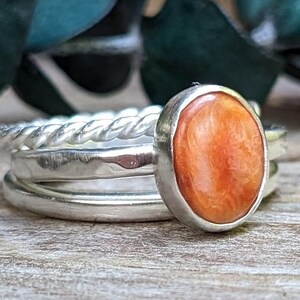 May include: A stack of silver rings, including a twisted band and two smooth bands. The top ring features an oval, orange gemstone set in a silver bezel. The rings are displayed on a wooden surface.