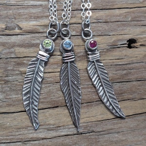 May include: Three silver feather pendant necklaces with different colored gemstones: green, blue, and red. The feathers have detailed veining and are attached to silver chains. The pendants are displayed on a rustic wooden surface.