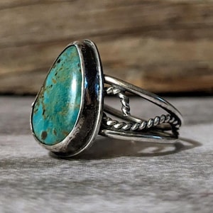May include: A silver ring with a teardrop-shaped turquoise stone. The stone is a vibrant teal color with brown matrix patterns. The ring has a twisted rope design on the band, adding a unique texture to the jewelry.