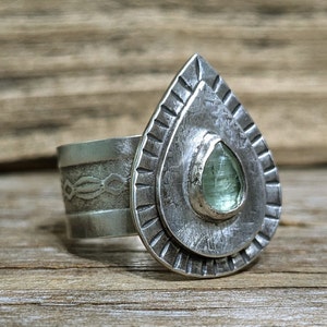 May include: A silver ring with a teardrop-shaped setting featuring a light green gemstone. The ring is decorated with intricate details and a textured finish.