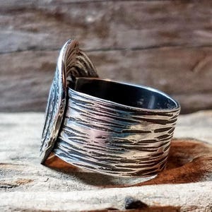 May include: A silver ring with a textured band and a decorative, leaf-like accent. The ring has a wide band with a rough, etched surface. The accent piece is a curved shape with detailed lines, adding a unique, artistic touch to the jewelry.