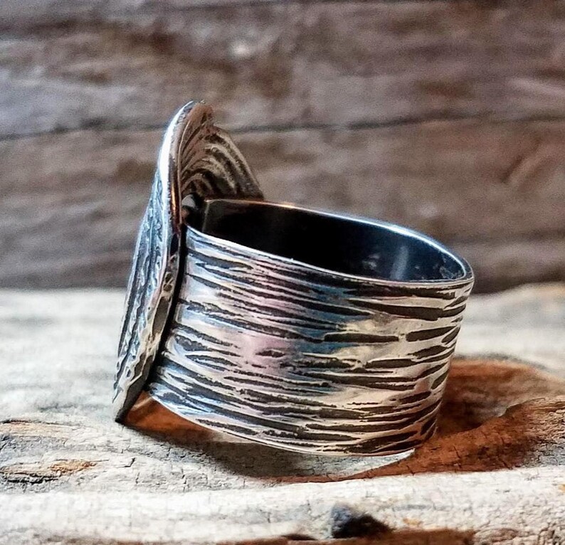May include: A silver ring with a textured band and a decorative, leaf-like accent. The ring has a wide band with a rough, etched surface. The accent piece is a curved shape with detailed lines, adding a unique, artistic touch to the jewelry.