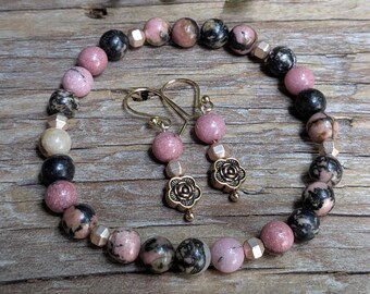 May include: A bracelet and earrings set featuring pink and black gemstone beads. The bracelet is strung with round beads and gold-colored spacers. The earrings have pink beads and gold-colored floral charms. The jewelry is displayed on a wooden surface.