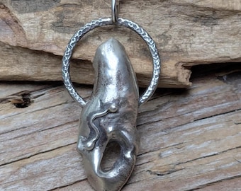 May include: A silver pendant necklace featuring a unique, abstract design. The pendant has an irregular shape with a textured surface and a small, decorative element. It hangs from a delicate silver chain. The pendant is approximately 2 inches long.