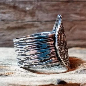 May include: A silver ring with a textured band and a decorative, leaf-shaped accent. The ring has a rustic, handcrafted appearance, with visible details in the metalwork. The ring is displayed on a wooden surface.