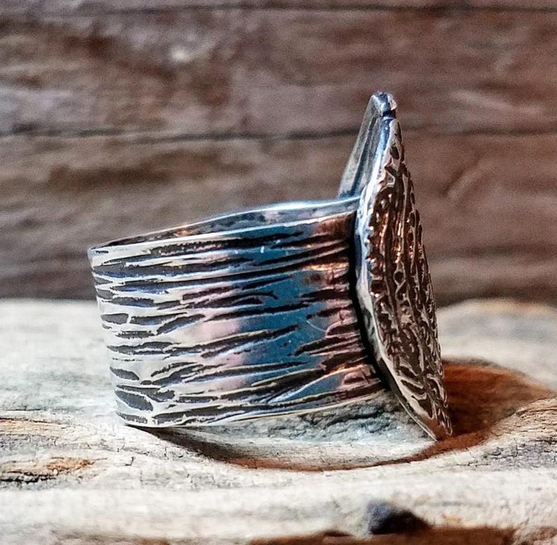 May include: A silver ring with a textured band and a decorative, leaf-shaped accent. The ring has a rustic, handcrafted appearance, with visible details in the metalwork. The ring is displayed on a wooden surface.