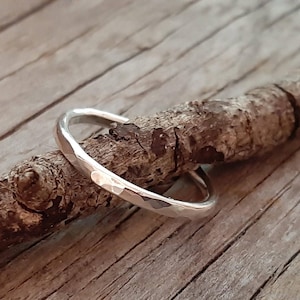 May include: A hammered silver ring with a textured surface. The ring is displayed on a small, brown wooden branch against a wooden background. The ring's design features a series of small, irregular facets that catch the light.