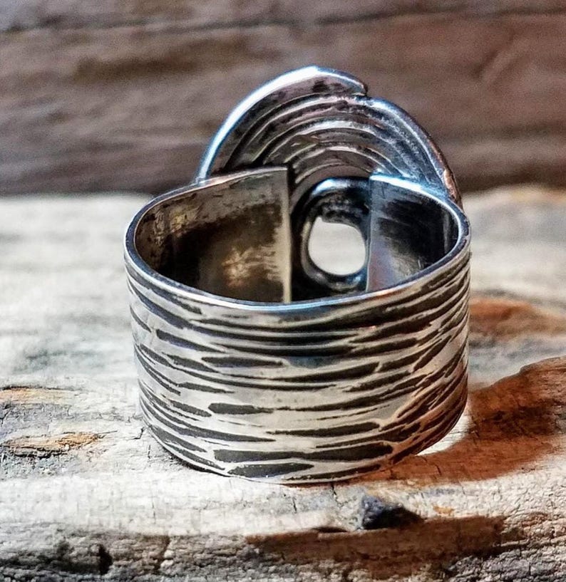 May include: A close-up of a silver ring with a textured band and a unique, layered design on top. The ring has a wide band with a textured, bark-like pattern. The top of the ring features a layered, curved design.