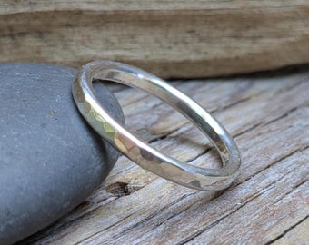 May include: A silver ring with a hammered texture. The ring is circular and sits on a gray stone, with a wooden background. The ring is a simple band design, reflecting light.