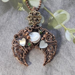 May include: A handcrafted copper wire pendant necklace. The pendant features a crescent moon design with a central iridescent stone, two heart-shaped accents, and small green beads. The necklace has a copper-colored chain.