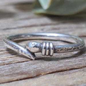 May include: A silver ring with a unique design. The ring features a textured band with a hook-like element and a hammered detail. The ring is set against a wooden surface.