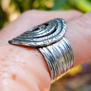 May include: A silver-toned ring with a textured band and a decorative, layered oval top. The oval top features a spiral design and intricate detailing. The ring is worn on a finger.