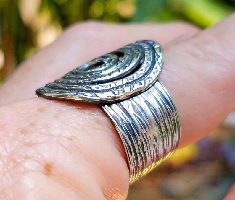 May include: A silver-toned ring with a textured band and a decorative, layered oval top. The oval top features a spiral design and intricate detailing. The ring is worn on a finger.