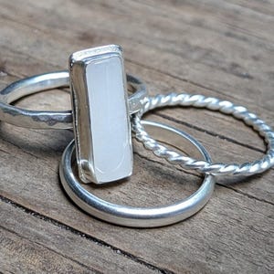 May include: A set of three silver rings featuring one ring with a vertical rectangular white stone, one ring with a twisted rope texture, and one plain smooth silver band. The rings are stacked together, showcasing different metal finishes.