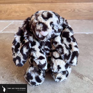 May include: A plush toy bunny with a leopard print pattern in shades of brown, black, and cream. The bunny has long, floppy ears, a small pink nose, and a soft, cuddly appearance. The toy is designed for children.