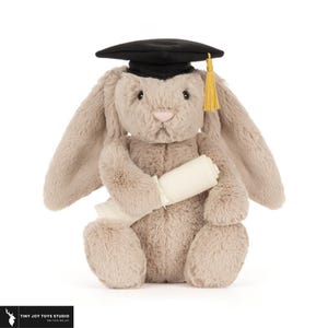 May include: A plush bunny wearing a black graduation cap with a gold tassel and holding a rolled-up scroll. The bunny is light brown with long ears and a pink nose, perfect for a graduation gift.