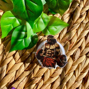 May include: A clear sticker showing a muscular figure lifting a barbell, wearing a crown of thorns. The figure is in brown, red, and white. The sticker is on a woven background with green leaves.