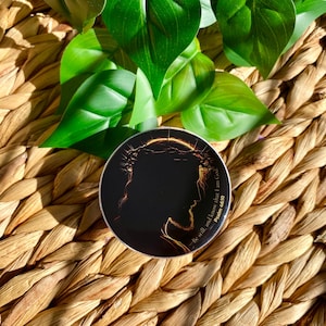 May include: A round, black religious-themed button with a gold outline of a person's profile wearing a crown of thorns. The text "Be still, and know that I am God. Psalm 46:10" is printed on the button. The button is on a woven surface with green leaves.