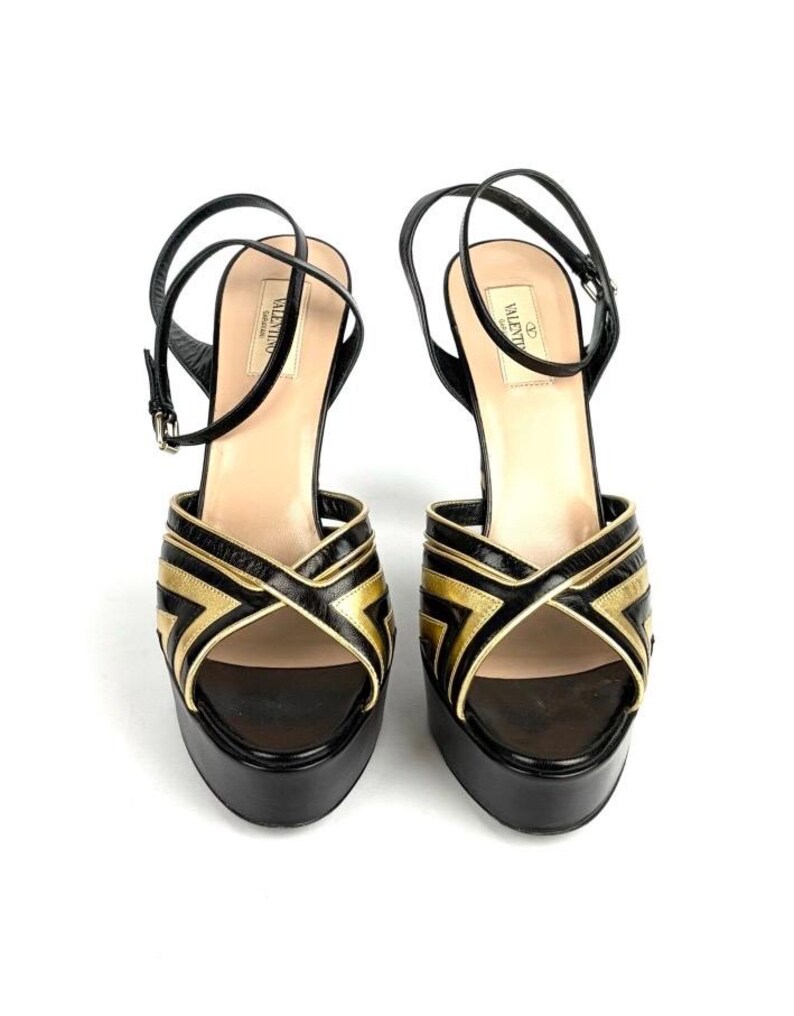 May include: A pair of black and gold platform sandals. The sandals feature a crisscross design on the front with gold accents. The ankle straps are black and the soles are black. The brand name "Valentino" is visible on the inner sole.