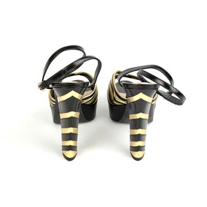 May include: Pair of black platform sandals with gold accents. The heels feature a black base with gold chevron and horizontal stripe designs. The sandals have a strap that buckles around the ankle.