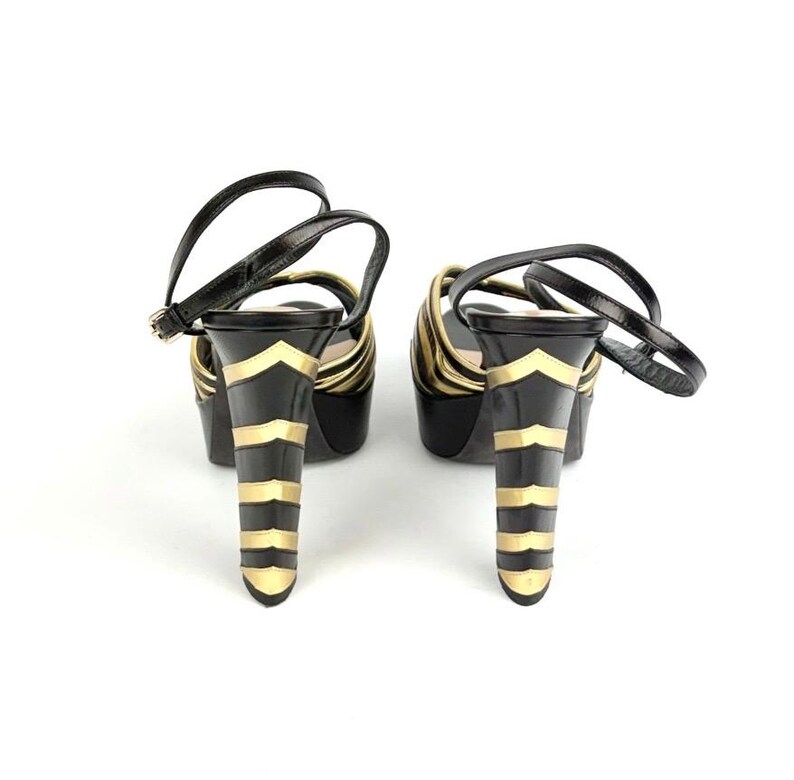 May include: Pair of black platform sandals with gold accents. The heels feature a black base with gold chevron and horizontal stripe designs. The sandals have a strap that buckles around the ankle.