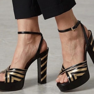 May include: Black and gold platform sandals with a thick heel. The sandals feature a crisscross design on the front and a thin ankle strap with a buckle. The heel has vertical gold stripes. The platform sole is approximately 1.5 inches high.