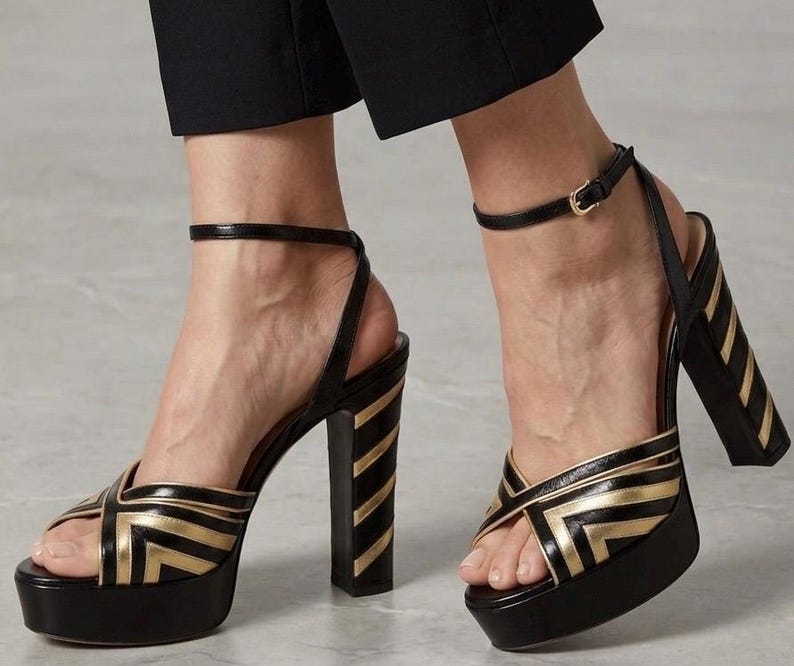 May include: Black and gold platform sandals with a thick heel. The sandals feature a crisscross design on the front and a thin ankle strap with a buckle. The heel has vertical gold stripes. The platform sole is approximately 1.5 inches high.