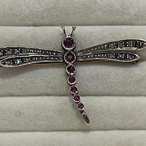 May include: A dragonfly brooch crafted from silver-toned metal, featuring a body set with a series of small, round, deep pinkish-red gemstones. The four wings are elongated and detailed with numerous tiny, sparkling clear stones throughout.