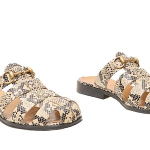 May include: A pair of slip-on sandals featuring a snakeskin pattern in shades of beige, cream, and black. The footwear has a cage-style upper with cutouts and a gold-toned metal horsebit detail across the top, set on a low, flat black heel.