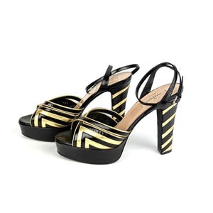 May include: A pair of black and gold platform sandals. The sandals feature a black base with gold chevron detailing on the straps and platform. The heels are striped in black and gold, and the ankle straps are black.