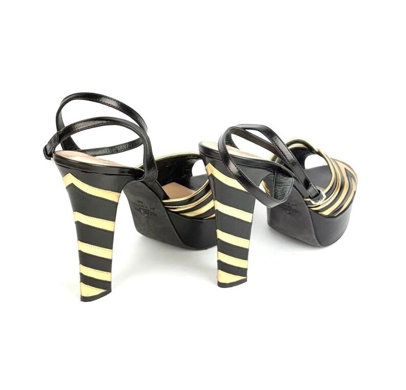 May include: Black and gold platform sandals with a striped design on the heels. The sandals have a black strap that wraps around the ankle and over the foot. The heels are approximately 5 inches tall.