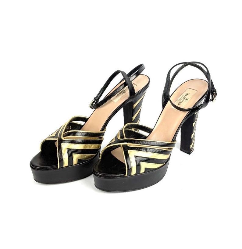 May include: Pair of black platform sandals with gold geometric accents. The sandals feature a thick platform sole, a thin ankle strap, and a high, striped heel. The brand name "Valentino" is visible on the inner sole.