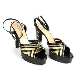 May include: Black and gold platform sandals with a geometric design. The sandals have a thick platform sole, a thin ankle strap, and a high block heel. The brand name "Valentino" is visible on the insole.