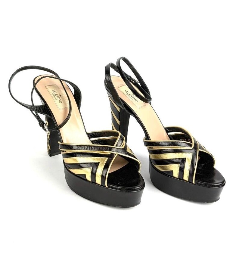 May include: Black and gold platform sandals with a geometric design. The sandals have a thick platform sole, a thin ankle strap, and a high block heel. The brand name "Valentino" is visible on the insole.