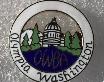 May include: A white enamel pin with a gold border. The pin features a white building with a dome, surrounded by green trees and a blue lake. The text "Olympia, Washington" is written around the outside of the pin, and the text "OWBA" is written in blue on the lake.