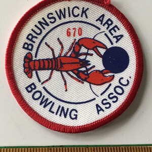 May include: A white circular patch with a red border. The patch has a blue circle with the text "Brunswick Area Bowling Assoc." in a blue arc around the circle. Inside the blue circle is a red lobster holding a blue bowling ball. The number "670" is printed in red above the lobster.