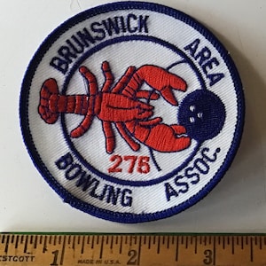 May include: A white, round patch with blue embroidery. The patch features a red lobster holding a blue bowling ball. The text "Brunswick Area Bowling Assoc. 278" is embroidered in blue around the lobster and bowling ball.