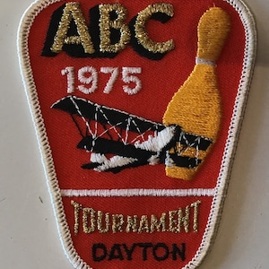 May include: A red, white, and gold embroidered patch with the text "ABC 1975 Tournament Dayton" and a black and white airplane with a yellow bowling pin.