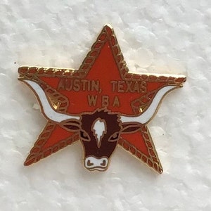 May include: A gold star-shaped pin with a red and white design. The pin features a brown longhorn steer with white horns and a white spot on its forehead. The text "Austin, Texas WBA" is printed on the star.