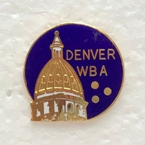 May include: A gold-coloured pin with a blue background. The pin features a white building with a gold dome and the text "DENVER WBA".