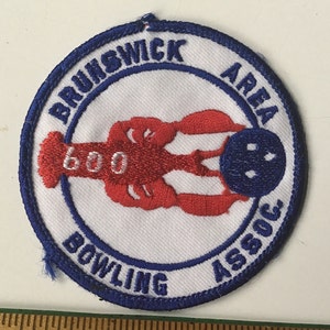 May include: A white and blue circular patch with the text "Brunswick Area Bowling Assoc." and a red lobster holding a blue bowling ball with white dots.