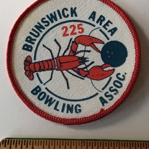 May include: A white round patch with a red border. The patch has a blue and white design with the text "Brunswick Area Bowling Assoc." and the number "225". A red lobster with a blue bowling ball is in the center of the patch.
