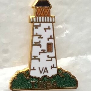 May include: A lighthouse pin badge featuring a white tower with gold edging and a black and gold top. The base is green with the letters "VA" and "TWBA". Likely a souvenir or collectible item.