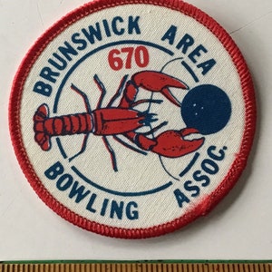 May include: A white, round patch with red stitching around the edge. The patch features a red lobster holding a blue bowling ball. The text "Brunswick Area Bowling Assoc." is written in a blue arc around the lobster. The number "670" is written in red above the lobster.