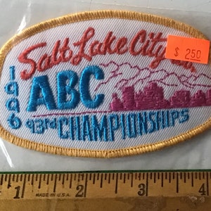 May include: Embroidered patch with the text "Salt Lake City ABC 1996 13th CHAMPIONSHIPS". The patch has a white background with red, blue, and pink lettering and a gold border. A price tag is attached.