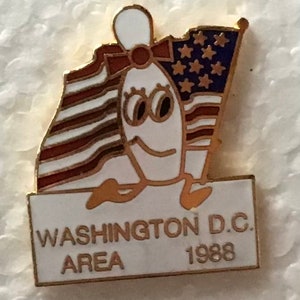 May include: A gold-coloured pin featuring a cartoon bowling pin wearing a red bow and holding an American flag. The pin is standing on a white rectangle with the text "WASHINGTON D.C. AREA 1988".