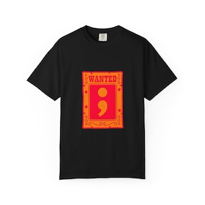 May include: Black t-shirt with a graphic design. The design features a red and orange frame with the word "WANTED" at the top. Inside the frame is a large orange semicolon. The shirt is made of a soft material.